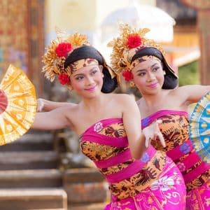 Dos mujeres con trajes tradicionales ornamentados y tocados dorados realizan un baile sincronizado sosteniendo abanicos coloridos.