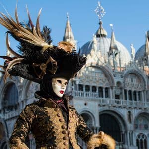 Un individuo con un elaborato costume di carnevale, maschera bianca e un grande cappello piumato, si erge di fronte a una basilica a cupola.