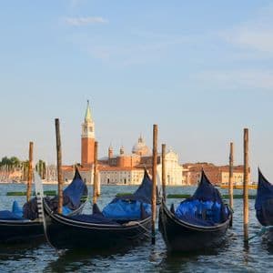 Diverse gondole con teloni blu sono ormeggiate a pali di legno in un canale, con un edificio storico e un campanile sullo sfondo.