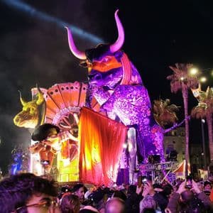 Un grande e sontuoso carro allegorico a forma di toro, illuminato da luci viola, si muove attraverso una folla fitta durante un festival notturno.