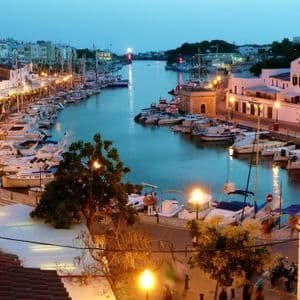 Ein abendlicher Blick auf einen Hafen voller festgemachter Boote, wo Ufergebäude und Straßenlaternen ein warmes Licht auf das Wasser zaubern.