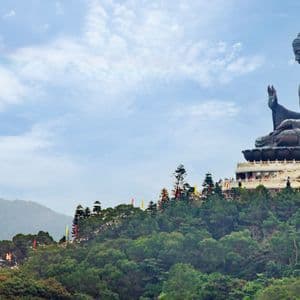 Une grande statue de Bouddha en bronze se dresse au sommet d'une colline boisée, sur une plate-forme à plusieurs niveaux, sous un ciel partiellement nuageux.