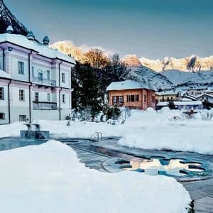 Una piscina termale esterna fumante in un villaggio innevato, con grandi montagne innevate sullo sfondo all'alba.