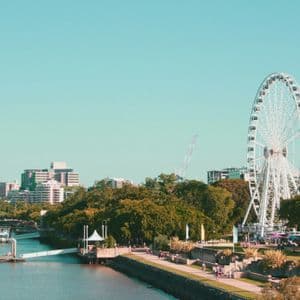 Une grande roue blanche se dresse sur les berges d'une ville, avec des gens se promenant sur une promenade bordée d'arbres verts luxuriants.