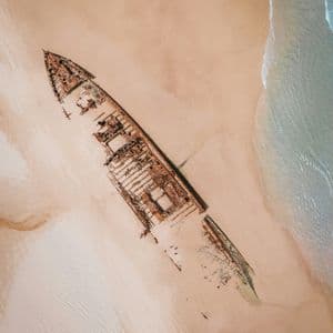 Vue aérienne d'une grande épave rouillée échouée sur le sable, avec de douces vagues de l'océan qui viennent s'échouer.