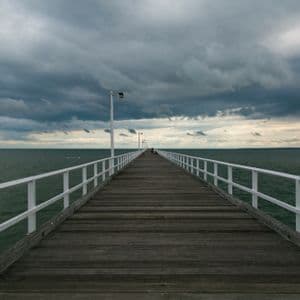 Une longue jetée en bois avec des balustrades blanches s'étend sur une mer sombre sous un ciel couvert et nuageux.