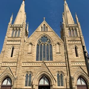 La façade d'une grande église en pierre avec deux flèches proéminentes, des arcs brisés et des vitraux sur un ciel bleu clair.