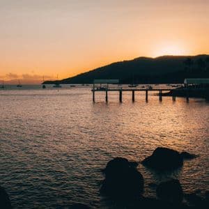 Le soleil se couche derrière une colline en silhouette, projetant une lueur orange sur une baie avec une jetée et des voiliers ancrés.