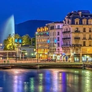 Una città su un fiume di notte con edifici illuminati, una grande fontana e una luna piena nel cielo.