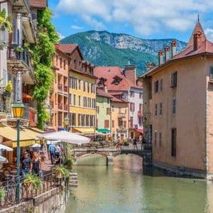 Un canale attraversa una città storica con edifici colorati e caffè all'aperto, con una grande montagna verde sullo sfondo sotto un cielo blu.