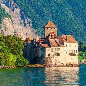 Un castello di pietra con tetti in tegole si trova sulla riva di un lago blu, con una montagna ripida e alberata sullo sfondo.