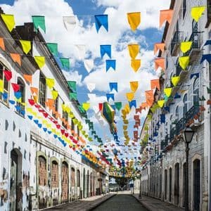 Colorful festival flags and a paper lantern hang over a narrow cobblestone street between old buildings under a partly cloudy sky.
