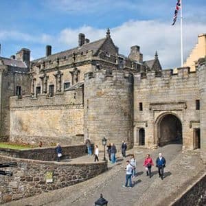 Varias personas caminan por un puente empedrado hacia la gran entrada de piedra de un castillo histórico bajo un cielo parcialmente nublado.