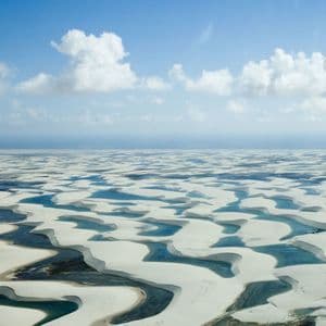 An aerial view of vast white sand dunes filled with lagoons of blue water under a partly cloudy sky.