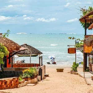 Ein sandiger Pfad zwischen rustikalen Gebäuden mit Reetdächern führt hinunter zu einem ruhigen Meeresstrand unter einem teils bewölkten Himmel.
