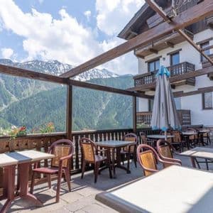 Una terrazza vuota di un ristorante all'aperto con tavoli e sedie, affacciata su una vista di montagne verdi e innevate.