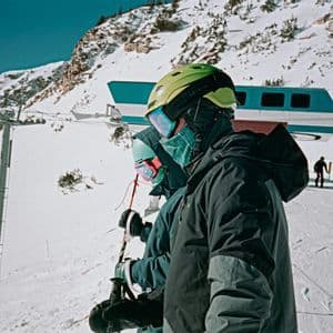 Un viaggio di gruppo WeRoad di sciatori con caschi e occhiali su una pista innevata, con una funivia e una seggiovia sullo sfondo.