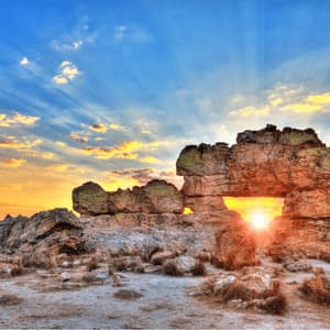 The setting sun shines through a natural arch in a large rock formation, with sunbeams radiating across a blue and yellow sky.