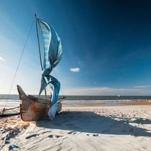 Ein Holzboot mit einem aufgerollten blau-weißen Segel ruht an einem Sandstrand neben dem Meer unter einem klaren blauen Himmel.