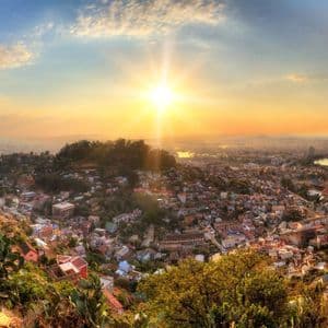A panoramic view of a city sprawling over hills, illuminated by a golden sunset with a prominent sunburst in the sky.