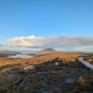 Une promenade en bois serpente à travers un paysage vallonné avec des lacs et des montagnes au loin sous un ciel partiellement nuageux.
