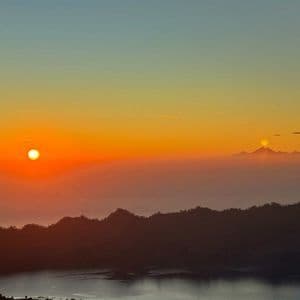 Una vivace alba crea un cielo sfumato su silhouette montuose stratificate e nebbia, con un secondo sole che emerge da una cima distante.