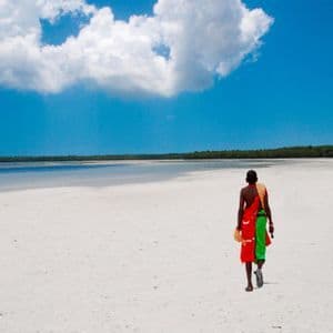 Une personne en tenue traditionnelle colorée traverse une vaste plage de sable blanc sous un ciel bleu avec des nuages moelleux.
