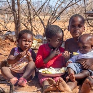 Quatre jeunes enfants sont assis par terre en extérieur et partagent un repas dans une seule assiette, l'un d'eux nourrissant un bébé.