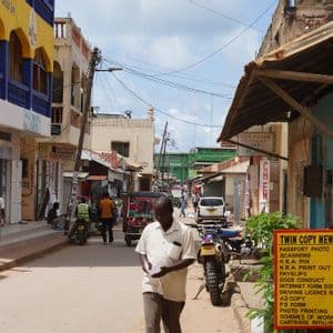 Un homme marche dans une rue animée bordée de bâtiments colorés, de boutiques et de motos sous un ciel partiellement nuageux.