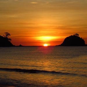 Il sole tramonta all'orizzonte tra due isole in silhouette, proiettando un bagliore arancione sul mare calmo.