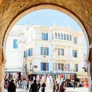 Una vista attraverso un grande arco di pietra di una soleggiata piazza pubblica con una fontana, bancarelle del mercato e un edificio bianco con persiane blu.