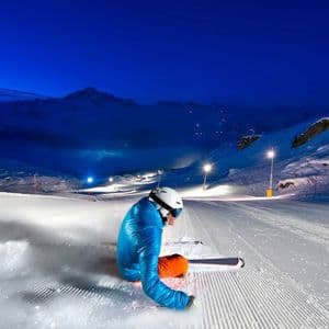 Uno sciatore con giacca blu e casco bianco siede su una pista da sci battuta di notte, illuminato da luci sotto un cielo scuro e stellato.