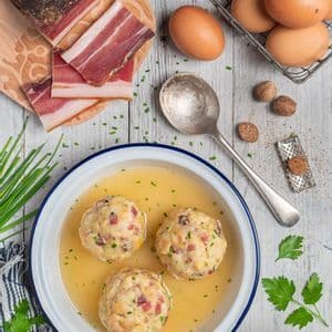 Una ciotola di canederli di speck in brodo su un tavolo di legno, circondata da ingredienti tra cui speck a fette, uova e cubetti di pane.