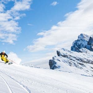 Uno sciatore in tuta gialla e nera scende una pista innevata, con cime montuose frastagliate sullo sfondo sotto un cielo parzialmente nuvoloso.
