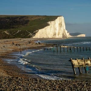 Imponenti scogliere bianche si incontrano con una spiaggia di ciottoli e un mare calmo, con persone che passeggiano lungo la riva sotto un cielo azzurro e limpido.