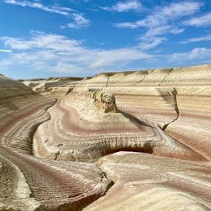 Colorful, layered rock formations of red and white sediment form rolling hills in a canyon under a blue sky.