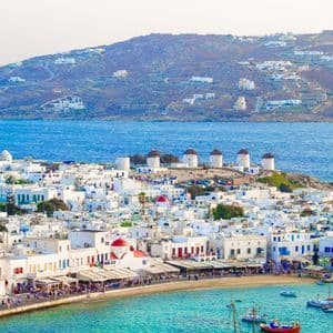 Un borgo costiero imbiancato a calce, con mulini a vento su una collina, si affaccia su una vivace baia turchese piena di piccole imbarcazioni.