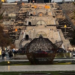 Una grande e intricata scultura in metallo a forma di teiera si trova in un parco, ai piedi di una monumentale scalinata in pietra a più livelli, al crepuscolo.