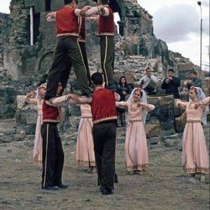 Un gruppo di persone in costumi tradizionali esegue una danza folcloristica, con gli uomini che formano una piramide umana davanti a rovine in pietra.