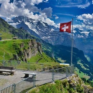 Un drapeau suisse sur un mât se dresse à un point de vue dominant une vallée verdoyante et une chaîne de montagnes enneigées.