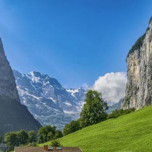 Une vue sur une vallée verdoyante encadrée par des falaises abruptes, avec des montagnes enneigées en arrière-plan sous un ciel bleu.