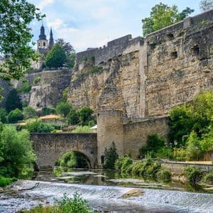 Un fiume con una chiusa scorre sotto un ponte ad arco in pietra alla base di grandi fortificazioni e una chiesa sulla collina.