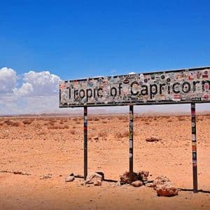 Un grande cartello con la scritta 'Tropico del Capricorno', coperto di adesivi, si erge in un vasto paesaggio desertico sotto un cielo blu.
