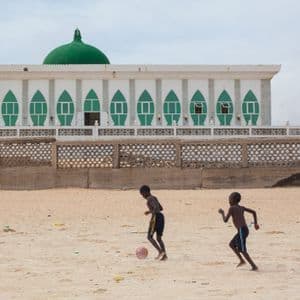 Due giovani ragazzi giocano a calcio su un terreno sabbioso davanti a una grande moschea bianca e verde con una cupola.