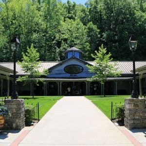 Un chemin pavé mène à un bâtiment rustique en bois avec un panneau de bienvenue, flanqué de pelouses vertes et d'une forêt dense.