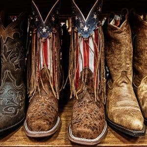 Diversi paia di stivali da cowboy in pelle con vari design allineati su uno scaffale, un paio con un motivo a bandiera americana.