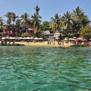Una vista dall'acqua limpida e turchese di una spiaggia sabbiosa affollata di persone sotto gli ombrelloni, con palme e edifici sullo sfondo.