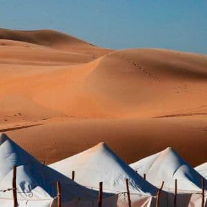 Una fila di tende bianche a tetto appuntito si trova alla base di grandi dune di sabbia arancioni sotto un cielo azzurro e limpido.