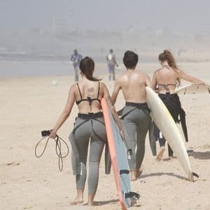 Un viaggio di gruppo WeRoad con tre persone in mute che camminano su una spiaggia sabbiosa con le loro tavole da surf verso l'acqua.