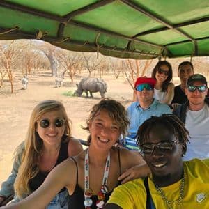 Un gruppo WeRoad in posa per una foto su un veicolo da safari mentre un rinoceronte e le zebre pascolano sullo sfondo.